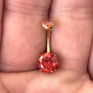 Puede incluir: Un piercing para el ombligo de color dorado con una gema roja grande y redonda en la parte inferior y una gema más pequeña a juego en la parte superior. La joya se sostiene en la palma de una mano abierta.