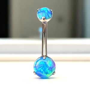 Può includere: Un piercing all'ombelico con una barra color argento e due sfere blu simili all'opale. Gli opali presentano una miscela di tonalità blu e verdi. Il gioiello è posto su una superficie bianca.