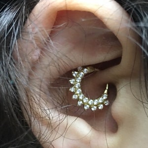 May include: A close-up of a person's ear with a gold hoop earring with small clear stones. The earring is in the upper part of the ear, and there is a small butterfly-shaped earring in the lower part of the ear.