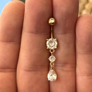Puede incluir: Anillo de ombligo dorado con tres piedras de circonita cúbica transparentes y brillantes. La piedra superior es redonda, la piedra central es más pequeña y redonda, y la piedra inferior tiene forma de pera. Joyería para piercing.