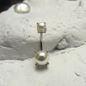 Puede incluir: Un piercing para el ombligo plateado con dos adornos de perlas falsas. La perla superior es cuadrada y la inferior es redonda. El anillo está hecho de metal brillante de color plateado.