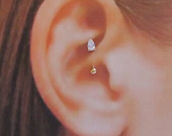 May include: A close-up of a person's ear with a clear teardrop-shaped gemstone and a small gold ball in a cartilage piercing.