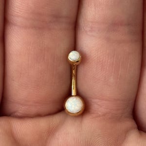 May include: A gold-colored belly button ring with two white opal-like stones. The curved barbell design features a small opal at the top and a larger one at the bottom. The jewelry is shown against a blurred background.