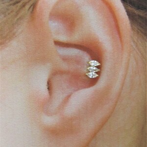 May include: A close-up of a person's ear with a small, gold, three-stone earring. The earring has three clear, round stones set in a row.
