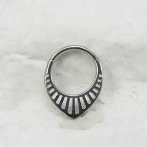 Könnte beinhalten: Ein silberfarbener, runder, segmentierter, offener Ring mit einer spitzen Unterseite. Der Ring hat ein dekoratives Design aus vertikalen Linien.