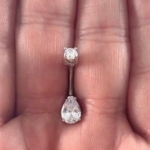 Puede incluir: Un piercing de ombligo plateado con una piedra transparente en forma de pera y una piedra redonda más pequeña.