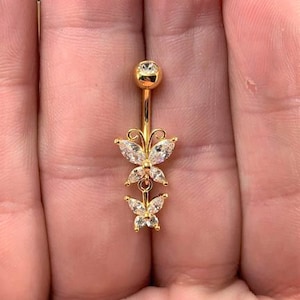May include: Gold-coloured belly button ring with a dangling butterfly design. The ring has a curved barbell with a clear gem at the top and two butterfly charms with clear gems. The butterflies are connected by a small chain.