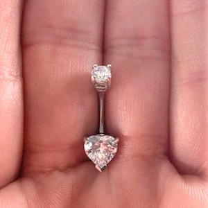 Puede incluir: Un piercing de ombligo plateado con una gema transparente en forma de corazón y una gema redonda transparente más pequeña.