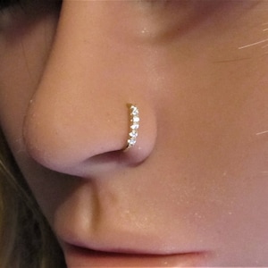 May include: A close-up of a person's nose with a small, gold nose ring set with clear cubic zirconia stones.