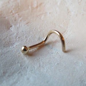 May include: A gold nose ring with a small ball at the end. The ring is curved and has a simple design.