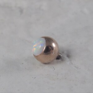 May include: A gold-toned body jewelry ball with a white opal top.