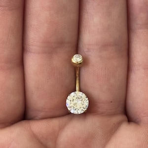May include: A gold-colored belly button ring with two clear, round, sparkling gemstones. The larger gemstone is at the bottom, and a smaller one is at the top. The ring is held in a person's hand.