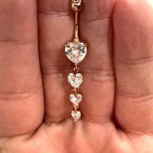 Könnte beinhalten: Ein goldener Bauchnabelring mit einer herabhängenden Kette aus fünf klaren herzförmigen Kristallen.