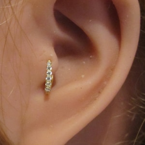 May include: A close-up of a person's ear with a small gold hoop earring with clear cubic zirconia stones.