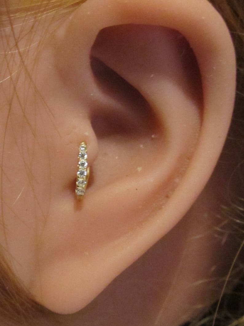 May include: A close-up of a person's ear with a small gold hoop earring with clear cubic zirconia stones.