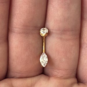 May include: A gold-coloured belly button ring featuring two clear, sparkling gemstones. One gemstone is round, and the other is an oval marquise shape. The ring is held in a hand.