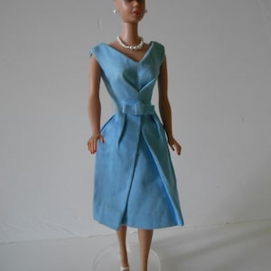 May include: A vintage doll in a light blue dress with a V-neck, a matching belt, and a pleated skirt. The doll has a blue headband, pearl earrings, and a pearl necklace. She is wearing white heels.