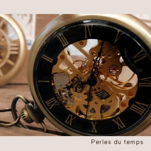 May include: A gold-toned pocket watch with a black face and Roman numeral markings. The watch has a skeleton design, revealing the intricate inner workings of the clock. The watch face also includes the text "Perles du temps".
