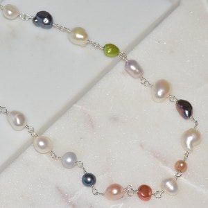 May include: A delicate necklace featuring a variety of freshwater pearls in different colors and sizes, including white, pink, black, and green. The pearls are strung on a silver chain.