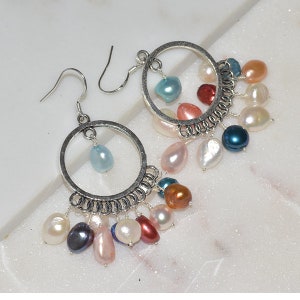 May include: Silver hoop earrings with a dangling chain of colorful pearls. The pearls are in shades of white, pink, blue, brown, and black.