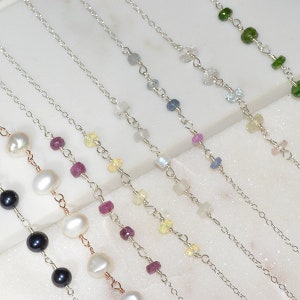 May include: Close-up of several delicate necklaces with silver chains and various gemstone beads, including pearls, rubies, and sapphires. The necklaces are displayed on a white surface, showcasing their colours and designs.