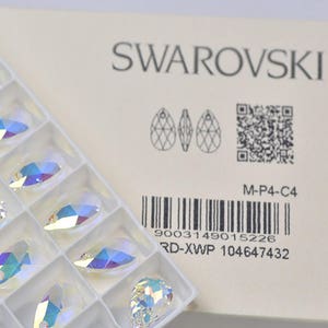 May include: A white box with the Swarovski logo and a barcode. The box contains a clear plastic tray with 12 crystal teardrop pendants.