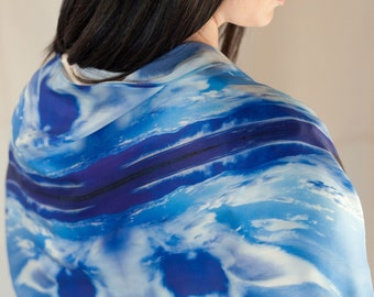 May include: A blue and white patterned scarf draped over the shoulders. The scarf features a watercolor-like design with shades of blue and white, creating a cloudy or ocean-like effect. The scarf is made of a lightweight material.