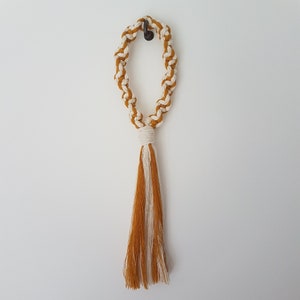 May include: A mustard yellow and white macrame tassel hanging from a hook. The tassel is made of braided cord and has a long, flowing fringe.