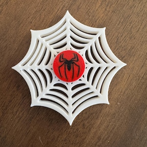 May include: A white, web-shaped decorative item with a red circle at the center. A black spider graphic is in the center of the red circle. The item is likely a decorative piece or emblem.