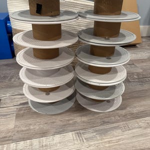 May include: Two stacks of white and gray tiered serving trays with a perforated design, each supported by a brown cardboard cylinder. The trays are arranged on a light wood-look floor, with a woven basket and blue object in the background.