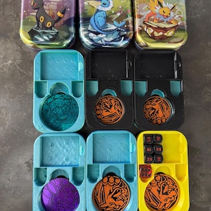 Könnte beinhalten: Eine Sammlung von Pokémon Trading Card Game Zubehör. Enthält drei Metalldosen mit Pokémon-Artwork und mehrere Kartenhalter aus Kunststoff mit Münzen und Würfeln. Die Dosen zeigen Umbreon, Vaporeon und Jolteon.
