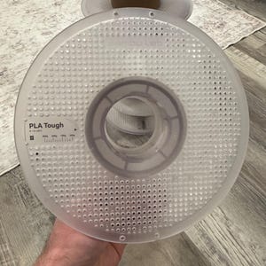 May include: A clear plastic spool of PLA Tough filament, with the text "PLA Tough" visible. The spool has a perforated design and is held by a person. Additional spools are stacked in the background.