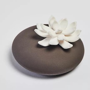 May include: A dark grey ceramic diffuser with a white ceramic flower on top. The flower has multiple petals and is in the shape of a lotus.