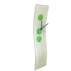 May include: A white, curved glass wall clock with black hands and three green circles. The clock has a modern design.