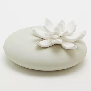 May include: A white ceramic diffuser with a white ceramic flower on top. The diffuser is shaped like a smooth, rounded stone.