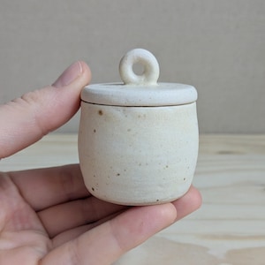 以下が含まれることがあります： 斑点模様の仕上げが施された、オフホワイトの小さな陶器製ジャーと蓋。蓋には中央に穴の開いた目立つ丸い取っ手が付いています。容器は滑らかな円筒形で、側面はわずかにカーブしており、マットな質感に仕上がっています。