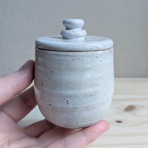 May include: A small, off-white ceramic jar with a matching lid. The jar features a speckled finish with brown flecks and horizontal ridges along the body. The lid has a rounded, tiered knob handle at the top, completing the cylindrical vessel.
