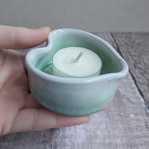 May include: A small, heart-shaped ceramic tealight candle holder in shades of pale green and white. The holder contains a white tealight candle with a visible wick. The holder is held in a hand.