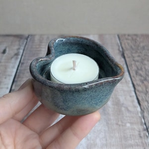May include: A heart-shaped teal ceramic candle holder with a white tea light candle inside. The candle holder has a glossy finish and a brown rim. The holder is being held by a person.