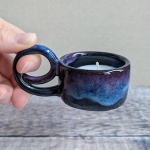 May include: A small ceramic candle holder with a circular handle, featuring a glossy glaze in shades of deep blue, purple, and black. The cylindrical vessel contains a white wax candle with a single centred wick, showcasing a smooth finish.