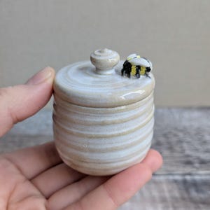 May include: A small, round, off-white ceramic jar with a matching lid. The jar features horizontal ridges and a glossy finish. A small, sculpted bee with black, yellow, and white details is attached to the top of the lid next to the knob.