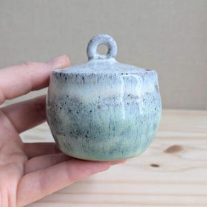 May include: A small, round ceramic jar with a lid featuring a circular handle. The piece has a speckled glaze that transitions from a pale, off-white top to a soft, muted green base, with dark brown flecks distributed throughout the surface.