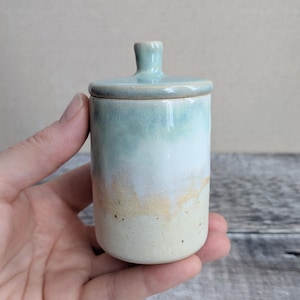 May include: A small cylindrical ceramic jar with a matching lid featuring a glossy glaze that transitions from a soft sage green at the top to a creamy off-white at the base, with subtle golden-brown speckles and a smooth, rounded finish.