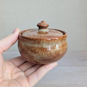 May include: A small, round ceramic jar with a lid. The jar is a warm brown colour with lighter, tan-coloured bands around the body. The lid has a small, rounded knob on top. The jar is held in a person's hand.