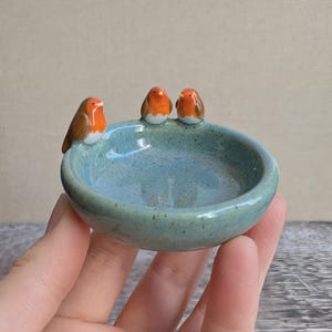 K&ouml;nnte beinhalten: Eine kleine, runde, hellblaue Keramikschale mit gesprenkelter Oberfl&auml;che. Drei Miniatur-Rotkehlchen-Figuren mit braunen Fl&uuml;geln, wei&szlig;er Brust und orangefarbenem Gesicht sitzen auf dem Rand der Schale und blicken zur Mitte des Gef&auml;&szlig;es.