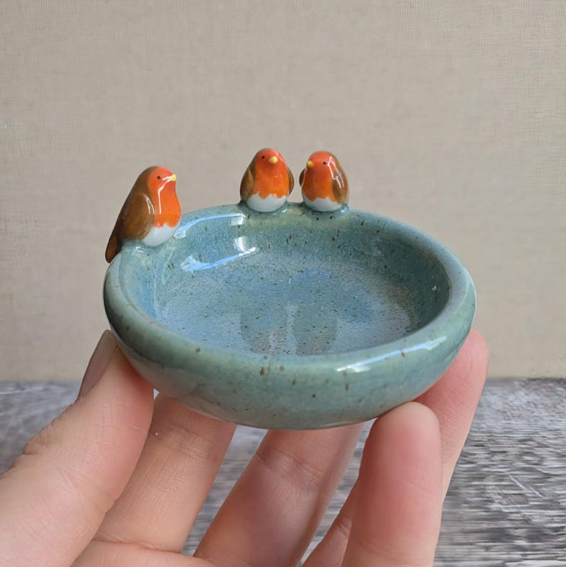 K&ouml;nnte beinhalten: Eine kleine, runde, hellblaue Keramikschale mit gesprenkelter Oberfl&auml;che. Drei Miniatur-Rotkehlchen-Figuren mit braunen Fl&uuml;geln, wei&szlig;er Brust und orangefarbenem Gesicht sitzen auf dem Rand der Schale und blicken zur Mitte des Gef&auml;&szlig;es.