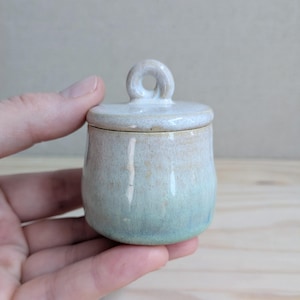 May include: A small, round ceramic jar with a matching lid featuring a circular handle. The vessel has a smooth, glossy glaze that transitions from a soft, pale cream colour at the top to a muted, light seafoam green hue at the base.