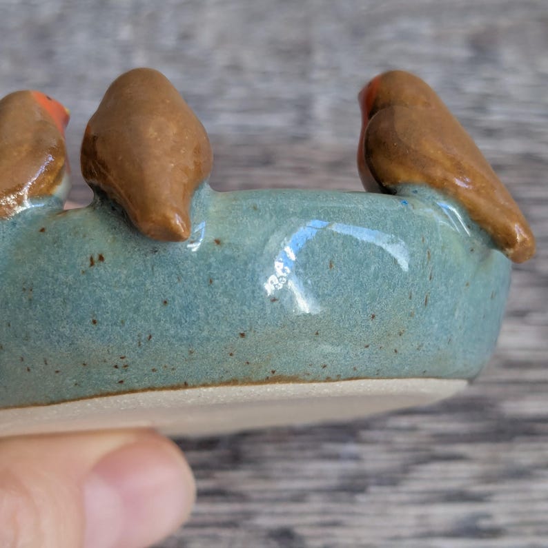 K&ouml;nnte beinhalten: Eine kreisf&ouml;rmige Keramikschale mit gesprenkelter blau-gr&uuml;ner Glasur, auf deren Rand drei kleine braune V&ouml;gel mit orangefarbenen Br&uuml;sten sitzen. Der Boden der Schale ist unglasiert und zeigt eine glatte, helle Tonoberfl&auml;che.