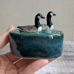 May include: A ceramic trinket box with a lid featuring two Canada geese figurines. The base is a deep, mottled teal and blue glaze, while the geese have black necks, white cheek patches, and brown and tan patterned bodies on the lid.