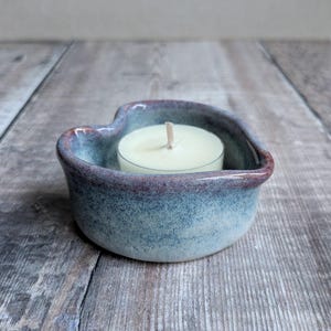 May include: A heart-shaped tealight candle holder with a blue and burgundy glaze. The ceramic holder contains a white tealight candle with a visible wick. The holder sits on a wooden surface.